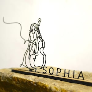 Könnte beinhalten: Eine schwarze Drahtskulptur einer Person, die Cello spielt. Die Skulptur ist auf einem Holzsockel mit dem Namen "SOPHIA" in schwarzen Buchstaben montiert. Die Skulptur steht auf einem hellbraunen Stein.