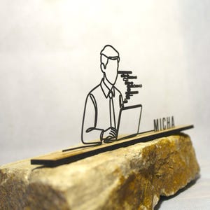 Op de afbeelding: Een zwart lijntekening bureau-accessoire met een persoon die op een laptop werkt. De naam "MICHA" staat op het accessoire. Het accessoire is geplaatst op een getextureerde, bruinachtige steen.
