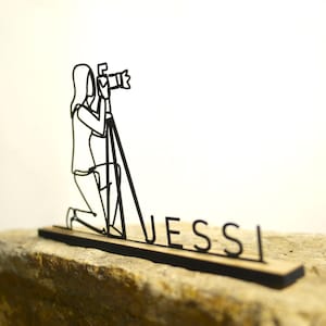 May include: Black metal wire sculpture of a photographer kneeling with a camera on a tripod. The sculpture is mounted on a wooden base with the name "Jessi" in black letters. The sculpture is set on a stone surface.
