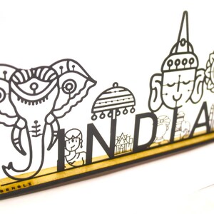May include: Black and white metal cutout of the word "INDIA" with various Indian symbols and designs, including an elephant, a person, an umbrella, and a temple. The cutout is mounted on a wooden base with the word "MADEHOLZ" printed on it.