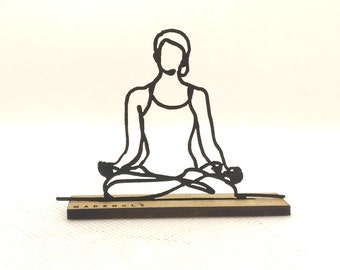 Könnte beinhalten: Schwarze Metall-Silhouette einer Yoga-Pose auf einem Holzsockel. Die Figur befindet sich in der sitzenden Lotusposition, Arme und Beine sind gekreuzt. Auf dem Sockel steht in Schwarz "MADEHOLZ". Der Hintergrund ist weiß.