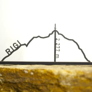 May include: A black metal silhouette of the Rigi mountain range, with the name "RIGI" and the height "2,277 m" displayed. The mountain outline is set against a white background and rests on a textured, golden-brown surface.