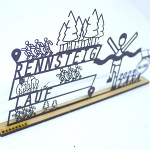 Könnte beinhalten: Schwarze Metallausschnitt einer Laufveranstaltung mit den Worten "Rennsteig Lauf" und einer Ziellinie mit einem Läufer, der sie überquert. Der Ausschnitt ist auf einem Holzsockel montiert.