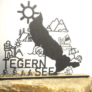 Op de afbeelding: Een zwart metalen decoratief bord met de omtrek van een meer en illustraties van buitenactiviteiten. Het bord bevat de tekst "TEGERN SEE" en afbeeldingen van fietsers, wandelaars en watersporten. Gemonteerd op een houten voet.