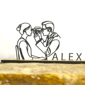 May include: A black laser-cut wooden desk accessory depicting an eye exam. The design shows a person examining another person's eyes. The name "ALEX" is displayed on the base. The accessory is placed on a stone surface.