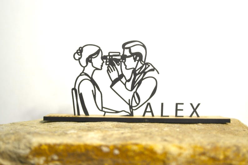 May include: A black laser-cut wooden desk accessory depicting an eye exam. The design shows a person examining another person's eyes. The name "ALEX" is displayed on the base. The accessory is placed on a stone surface.