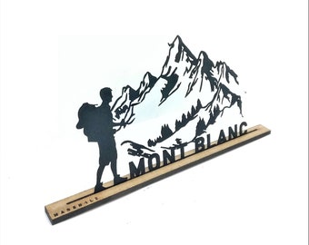 Op de afbeelding: Zwarte silhouet van een wandelaar met een rugzak die voor een bergketen staat met de tekst "MONT BLANC" op een houten basis.