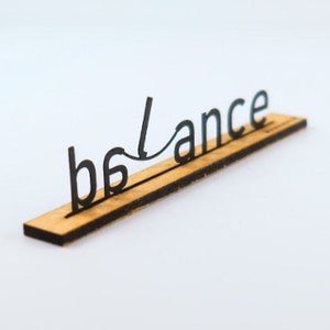 Könnte beinhalten: Ein Holzschild mit dem Wort "balance" in schwarzen Buchstaben. Die Buchstaben sind so angeordnet, dass das Schild ausgeglichen erscheint.