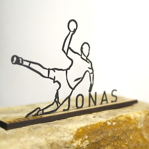 Puede incluir: Una ilustración en línea negra de un jugador de balonmano en acción, sosteniendo un balón. La silueta del jugador está sobre el nombre "JONAS" recortado de una base de madera. La obra de arte está sobre una superficie texturizada de color marrón claro.