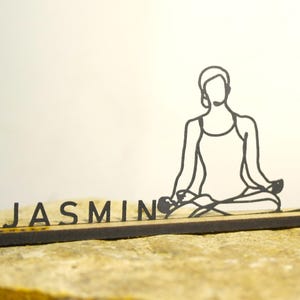 Könnte beinhalten: Schwarze, lasergeschnittene Yoga-Dekoration aus Holz. Das Design zeigt eine sitzende Figur in einer Yoga-Pose, mit dem Namen "JASMIN" davor. Die Figur und die Buchstaben befinden sich auf einem Holzsockel vor neutralem Hintergrund.