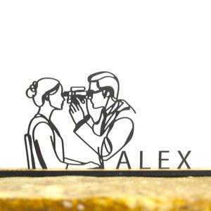 May include: A black and white line drawing of a person getting their eyes examined by an eye doctor. The name "ALEX" is displayed in black letters on a wooden base. The image is set against a white background.