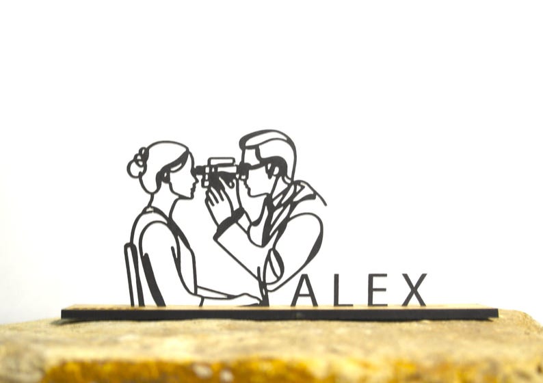 May include: A black and white line drawing of a person getting their eyes examined by an eye doctor. The name "ALEX" is displayed in black letters on a wooden base. The image is set against a white background.