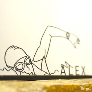 Puede incluir: Escultura de alambre negro de un nadador en estilo libre, con gafas y gorro de natación. El nombre "ALEX" se muestra en letras individuales sobre una base negra. Sobre un fondo blanco.