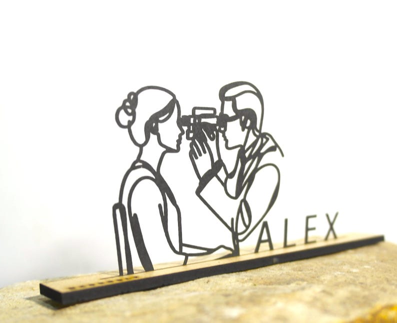 May include: A black laser-cut wooden decoration depicting a person looking through a camera at another person. The word "ALEX" is cut out below the figures. The figures are on a rectangular wooden base.