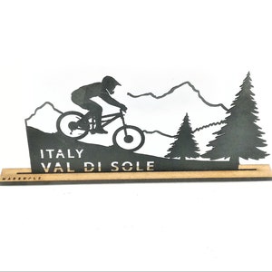 Könnte beinhalten: Schwarze Silhouette eines Mountainbikers, der einen Bergabhang in einer Berglandschaft hinunterfährt. Der Text "ITALY VAL DI SOLE" ist auf der Basis des Holzschildes gedruckt.