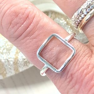 May include: A polished silver ring featuring a simple, open square-shaped design on the front. The band is thin and smooth, encircling the finger. An additional textured silver band is visible on the adjacent finger, completing the look.