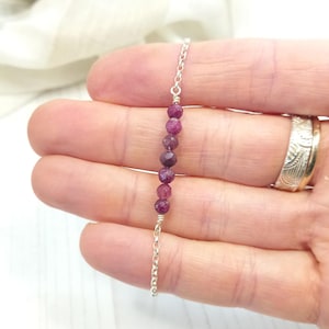 May include: A silver chain bracelet with a line of faceted, deep red ruby beads. The bracelet is held in a hand, highlighting the polished gemstones and the fine silver chain links. The bracelet is a piece of jewellery.
