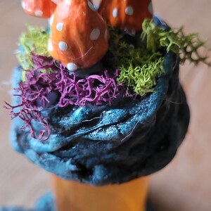 May include: A miniature scene with three orange and white mushrooms on a dark blue and green base with purple and green moss. The base is on a clear glass jar.