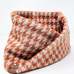 May include: A houndstooth patterned neck warmer in shades of orange and cream. The fabric is folded over, revealing the interior. A small, brown leather tag with a logo is attached to the bottom.
