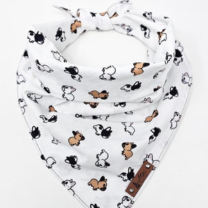 May include: White cotton bandana with a pattern of small black and brown dogs. The bandana has a brown leather tag with the text "The Doggy" on it.