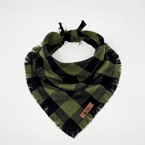 May include: A green and black plaid dog bandana with a fringed edge. The bandana is tied at the top and features a small brown leather tag with a logo. The fabric has a soft, textured appearance.