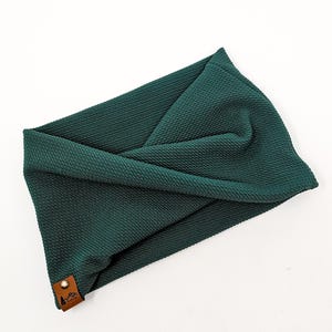Puede incluir: Una diadema retorcida de color verde oscuro hecha de tela texturizada. La diadema tiene una pequeña etiqueta de cuero marrón con un logotipo. La tela parece ser un material de punto o tejido.
