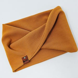 May include: Mustard-colored, textured fabric headband. The headband is folded and has a small, brown leather tag with a mountain and tree logo. The fabric has a woven pattern and is a warm, earthy tone.
