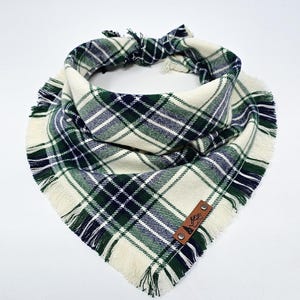 May include: A white, green, and blue plaid dog bandana with frayed edges. The bandana has a brown leather patch with the text "The Barkery" on it.