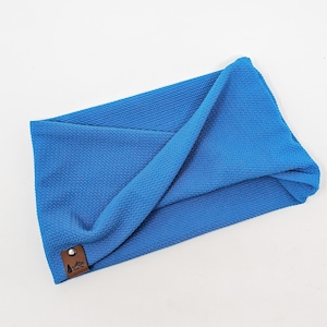 May include: A vibrant blue headband with a textured, woven pattern. The headband is folded over itself and has a small, brown leather tag with a mountain and tree logo. The accessory is designed to be worn around the head.
