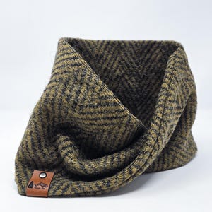 Puede incluir: Bufanda tubular con estampado de espiga en verde oliva y negro. La bufanda tiene una etiqueta de cuero marrón con el texto "Pametic Pine" y un gráfico de montaña y árbol. Hecha de un material suave y texturizado.