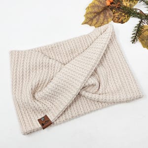 May include: A cream knitted headband with a brown leather tag. The headband is folded in half and has a subtle herringbone pattern.
