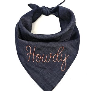 Puede incluir: Un pa&ntilde;uelo para perro de mezclilla azul marino con la parte superior anudada. La palabra "Howdy" est&aacute; bordada con hilo naranja claro en la parte delantera del pa&ntilde;uelo. El pa&ntilde;uelo es de forma triangular.
