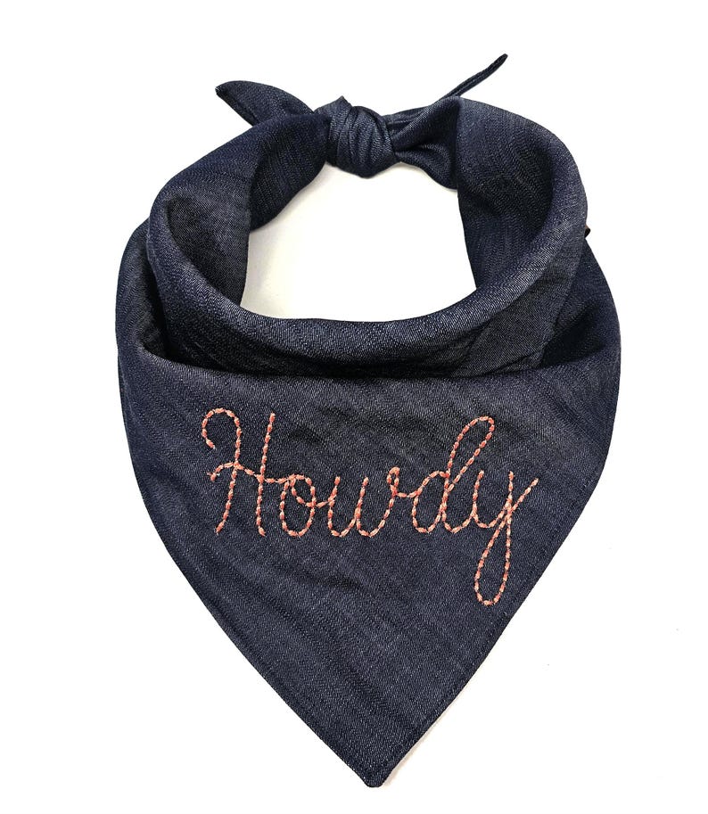 Puede incluir: Un pa&ntilde;uelo para perro de mezclilla azul marino con la parte superior anudada. La palabra "Howdy" est&aacute; bordada con hilo naranja claro en la parte delantera del pa&ntilde;uelo. El pa&ntilde;uelo es de forma triangular.