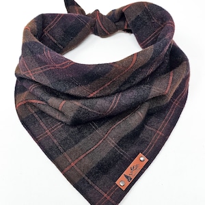 May include: A plaid dog bandana in shades of brown, black, and red. The fabric is knotted at the top and features a leather tag with a mountain and tree logo. The bandana is designed to be worn around a dog's neck.
