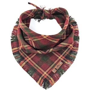 May include: A dog bandana in a red, green, and yellow plaid pattern. It features a fringed edge and a leather tag with a mountain and tree logo. The bandana is tied at the top.