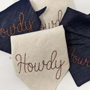 Puede incluir: Cuatro pa&ntilde;uelos para perros, dos azul marino y dos beige claro. Cada uno tiene la palabra "Howdy" bordada en hilo marr&oacute;n rojizo. Los pa&ntilde;uelos est&aacute;n doblados para mostrar el texto. Primer plano de los pa&ntilde;uelos.