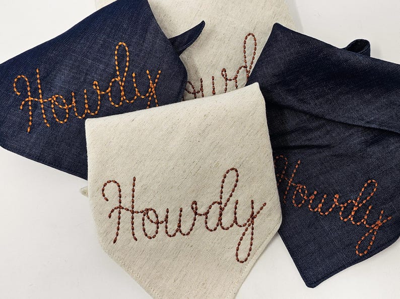 Puede incluir: Cuatro pa&ntilde;uelos para perros, dos azul marino y dos beige claro. Cada uno tiene la palabra "Howdy" bordada en hilo marr&oacute;n rojizo. Los pa&ntilde;uelos est&aacute;n doblados para mostrar el texto. Primer plano de los pa&ntilde;uelos.