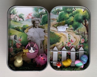 May include: A rectangular metal tin opened to reveal a miniature diorama featuring a vintage-style illustration of a child in a white dress, a rabbit in a pink basket, a yellow chick, a white sheep, a white picket fence, and colorful eggs.
