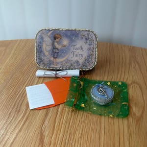 May include: A rectangular metal tin featuring a fairy illustration and the text Tooth Fairy, a small white scroll tied with silver string, a bright orange envelope, and a green organza pouch containing a blue circular charm with a silver fairy.