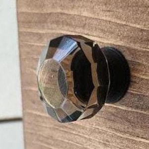 May include: A faceted, clear and black cabinet knob is mounted on a stained wooden surface. The knob has a geometric design and a black base. The wood grain is visible, adding texture to the background.