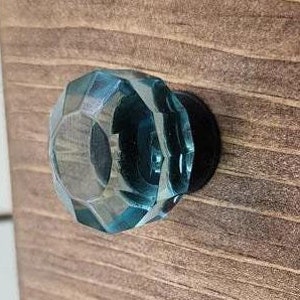 May include: A teal blue glass cabinet knob with a faceted design. The knob is mounted on a wooden cabinet door.