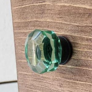 May include: A wooden cabinet with a faceted, light green glass knob. The knob is mounted on a black base. The wood has a natural grain and a warm brown stain.