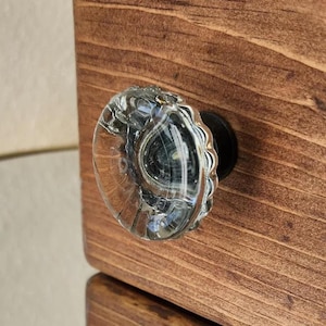 May include: A clear glass knob with a decorative, textured surface. The knob is attached to a wooden drawer or cabinet.