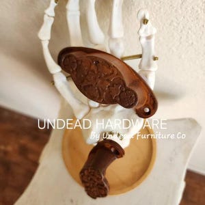 May include: Two rusty brown metal drawer pulls with ornate designs. The pulls are displayed on a skeleton hand. The text "UNDEAD HARDWARE By Undead Furniture Co." is visible below the pulls.