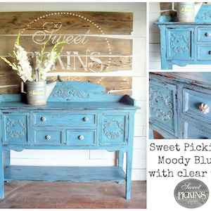 May include: A distressed blue painted sideboard with carved details and silver knobs. The sideboard has four drawers and a lower shelf. The sideboard is labeled "Sweet Pickins Moody Blue with clear wax".