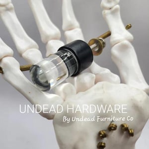 May include: A clear glass vial with a black cap, secured with a bolt and washer, is held by a white skeletal hand. The text "UNDEAD HARDWARE By Undead Furniture Co" is visible.