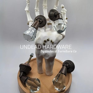 May include: Five antique-style cabinet knobs with clear glass crystal finials. The knobs are displayed on a wooden base and a skeletal hand. The text "UNDEAD HARDWARE By Undead Furniture Co" is visible below the hand.