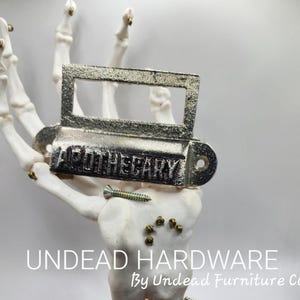 May include: Aged silver-toned metal apothecary sign held by a white skeleton hand. The sign reads "APOTHECARY" in embossed letters. A screw is placed below the sign. The text "UNDEAD HARDWARE By Undead Furniture Co" is at the bottom.