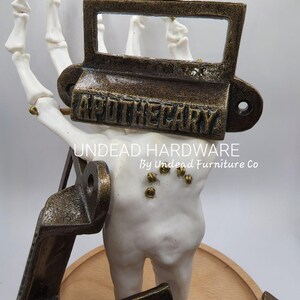 May include: Aged bronze-colored metal apothecary label holder with the word "APOTHECARY" embossed on it. The label holder is held by a white skeleton hand. The words "UNDEAD HARDWARE By Undead Furniture Co" are also visible.