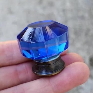 May include: A faceted, cobalt blue glass cabinet knob with a bronze-colored base. The knob has a geometric design and reflects light, creating a sparkling effect. This decorative hardware is suitable for furniture restoration or DIY projects.